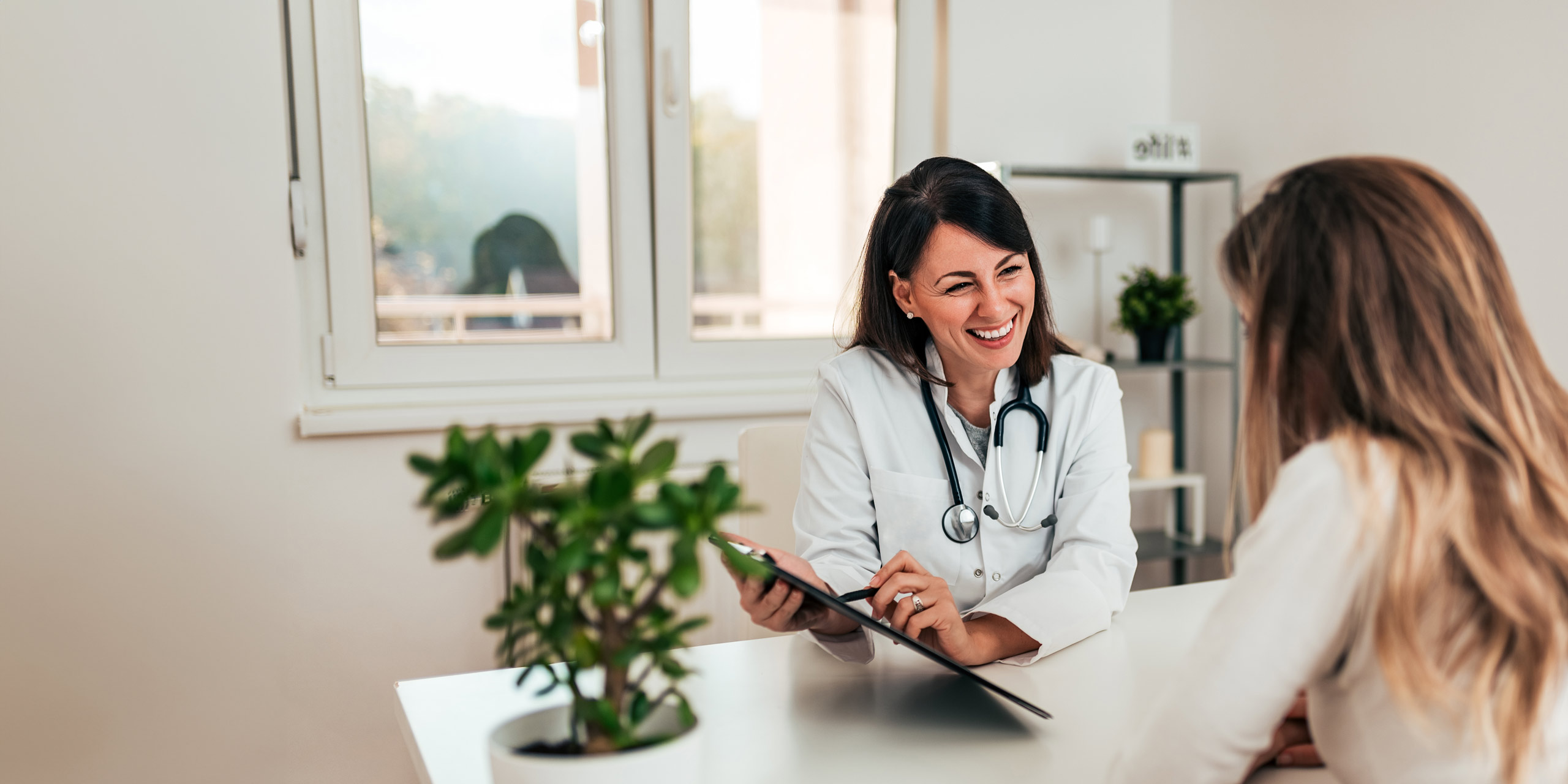 Eine Ärztin sitzt mit einem Stethoskop um den Hals an ihrem Schreibtisch, lächelt und hält ein Tablet in der Hand. Sie unterhält sich mit einer ihr gegenübersitzenden Frau. Der Raum hat große Fenster und im Vordergrund steht eine Topfpflanze.