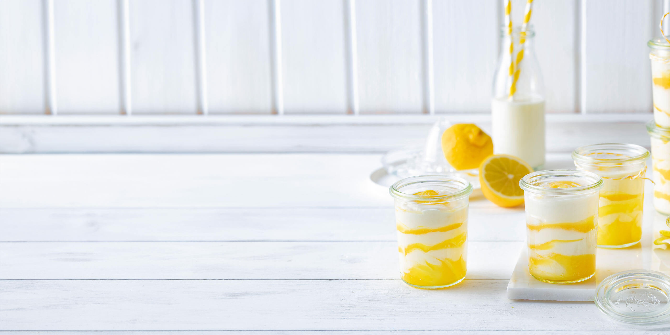 Gläser mit Lemon-Curd-Mousse mit frischen Zitronenscheiben auf einem weißen Holztisch, dazu eine Flasche Milch und eine ganze Zitrone. Helles, erfrischendes Ambiente.